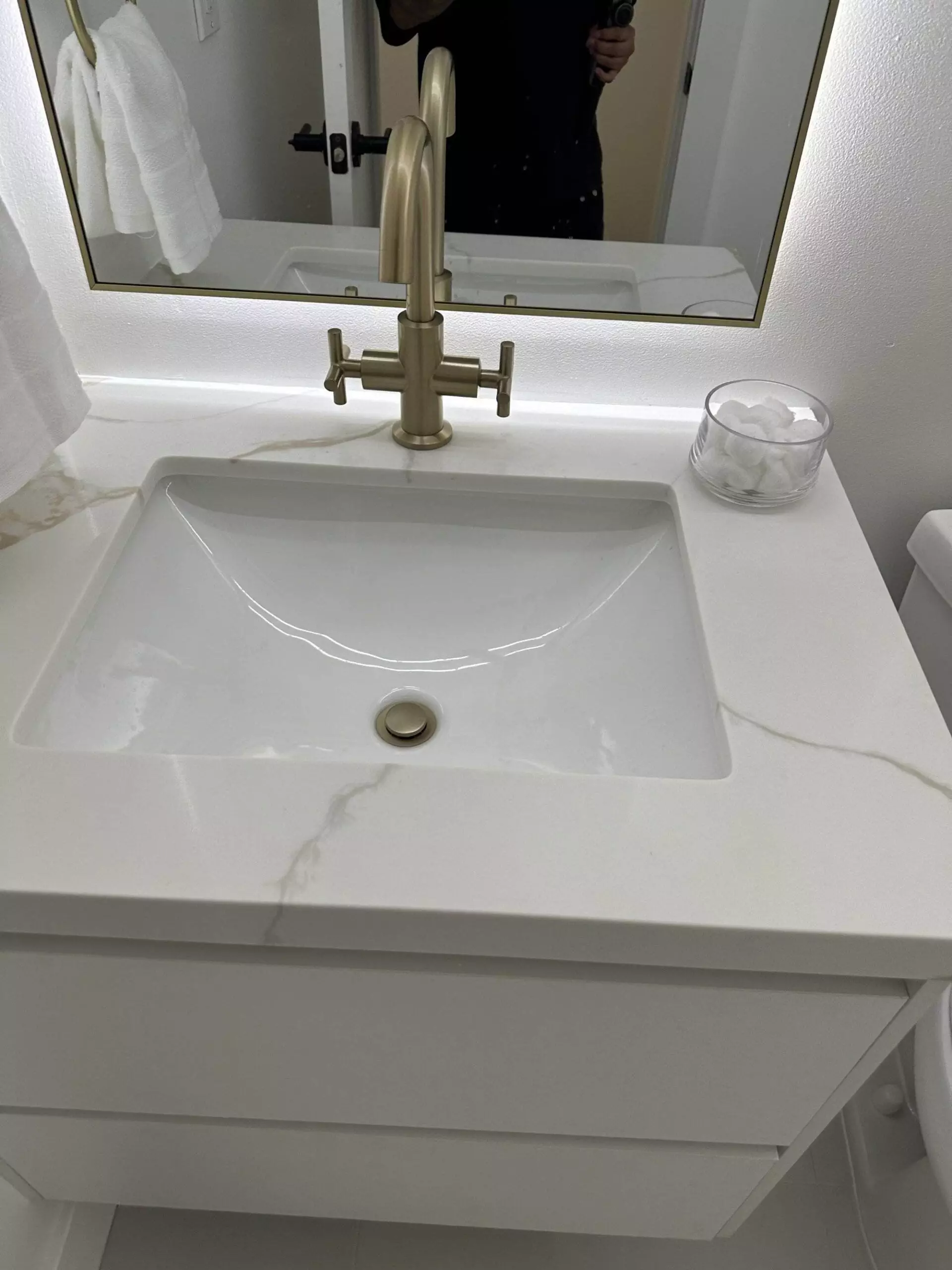 Modern bathroom sink with a gold faucet, white marble countertop, and a glass jar holding cotton balls, reflected in a mirror above.