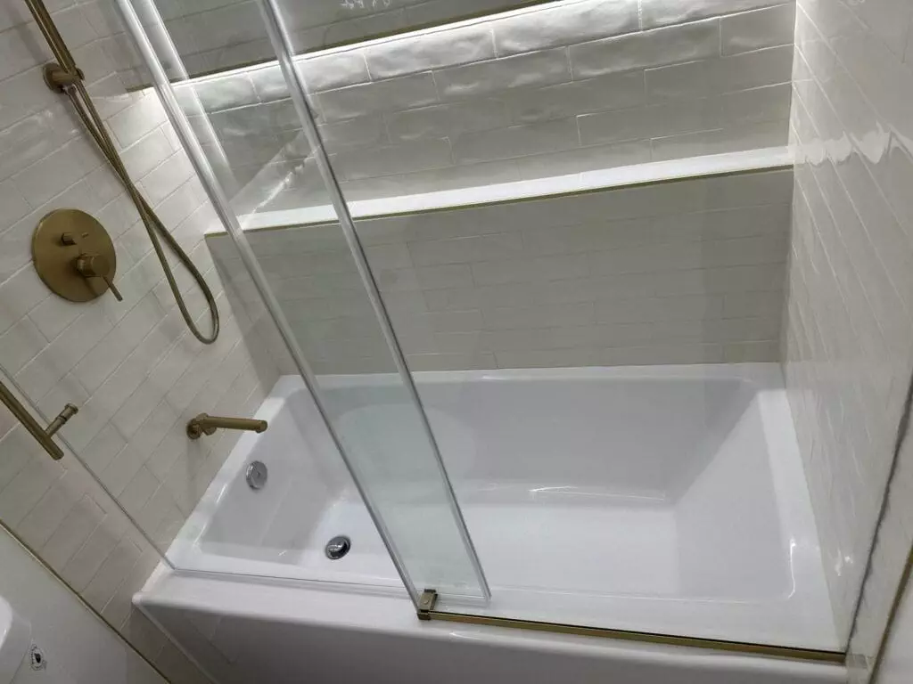 A clean, white bathtub with a sliding glass door, situated in a tiled bathroom with brass fixtures, and a handheld showerhead on the wall.