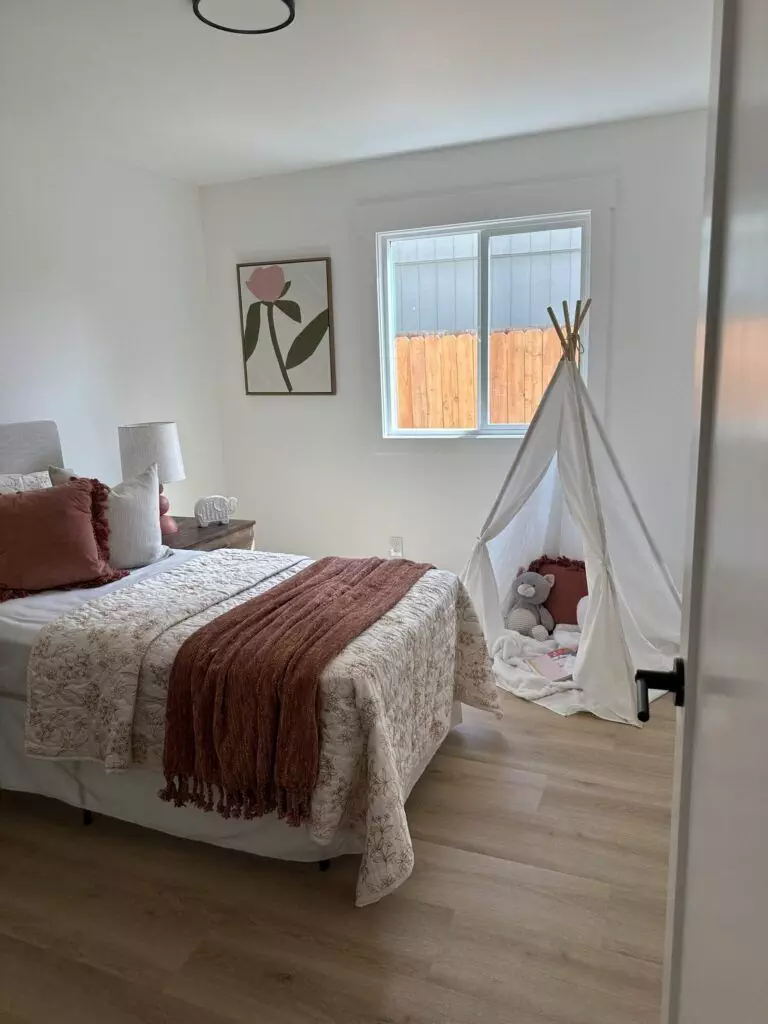 A small bedroom with a neatly made bed adorned with patterned bedding and a brown throw. A child's teepee tent with stuffed animals is set up near a window. A framed flower picture hangs on the wall.