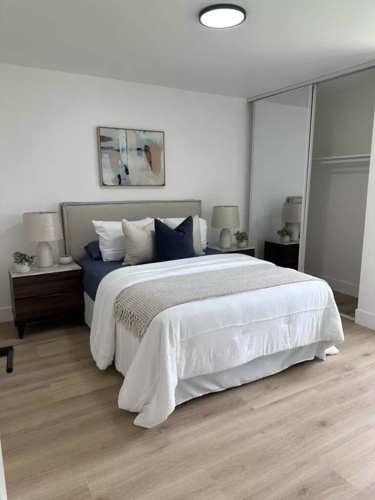 A neatly made bed with multiple pillows is centered between two matching nightstands with lamps. A painting hangs above the headboard, and a sliding mirror door closet is on the right. The floor is wooden.