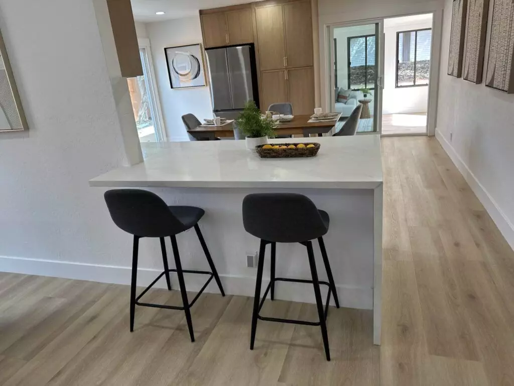 A kitchen with a white marble island, two gray barstools, a stainless steel refrigerator, and wooden cabinets. An open dining area with a table and chairs is visible in the background.