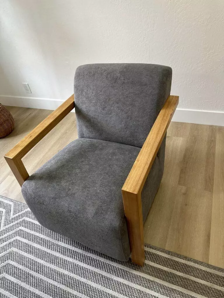 A modern armchair with gray upholstery and wooden armrests sits on a wooden floor next to a striped rug.