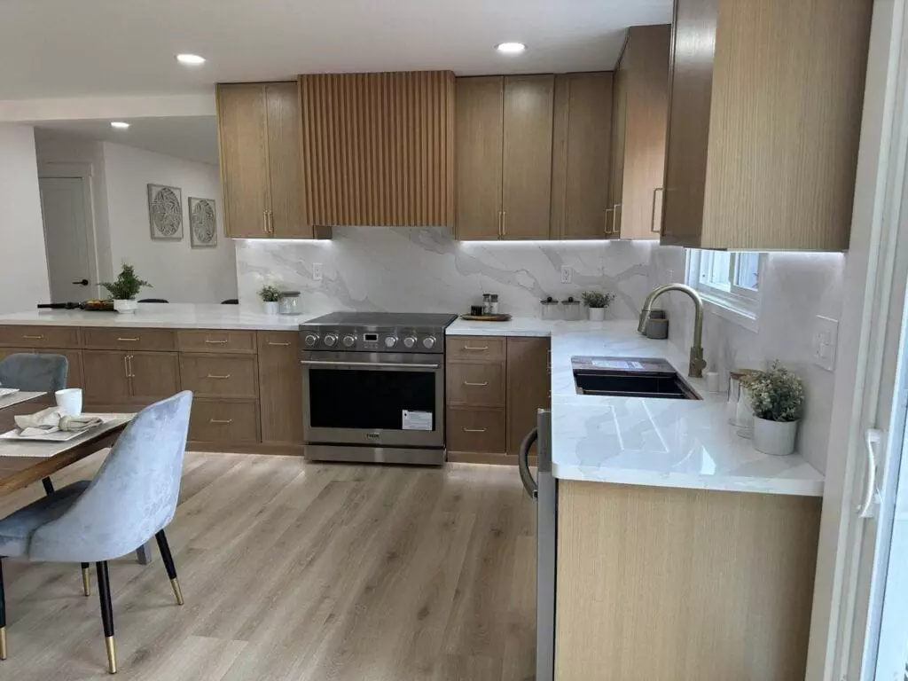 A modern kitchen with wooden cabinets, a stainless steel stove, marble countertops, and light wood flooring. A dining area with grey chairs and a wooden table is visible on the left.