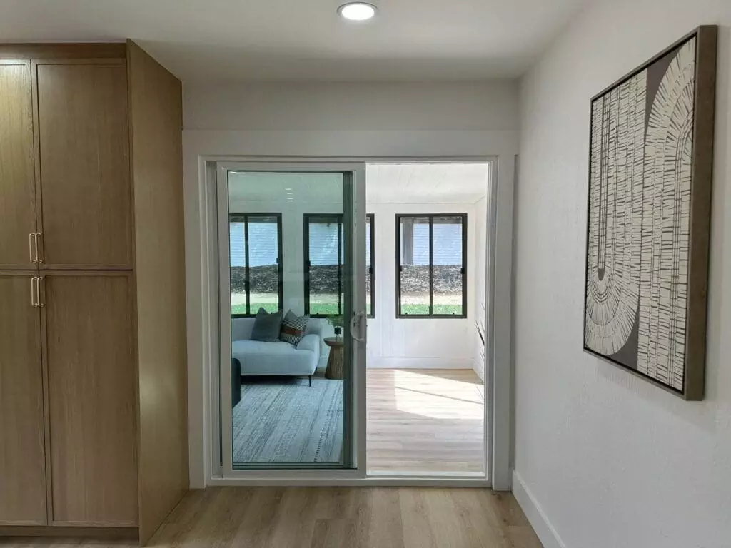 A wooden floor hallway with built-in cabinets on the left, a large abstract painting on the right, and a sliding glass door leading to a sunroom with a sofa and multiple windows.