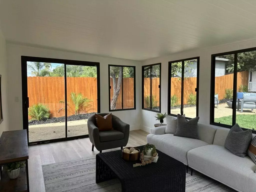 A bright, modern sunroom with large windows, a white sectional sofa, a black coffee table, and a gray armchair. Outside, there's a wooden fence with greenery along its base.