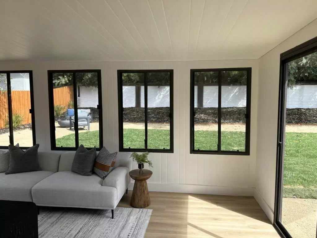 A bright indoor room with multiple large windows, a white ceiling, light wood flooring, a beige sectional sofa, and a small round wooden side table. Outside, there's a grassy backyard and a wooden fence.