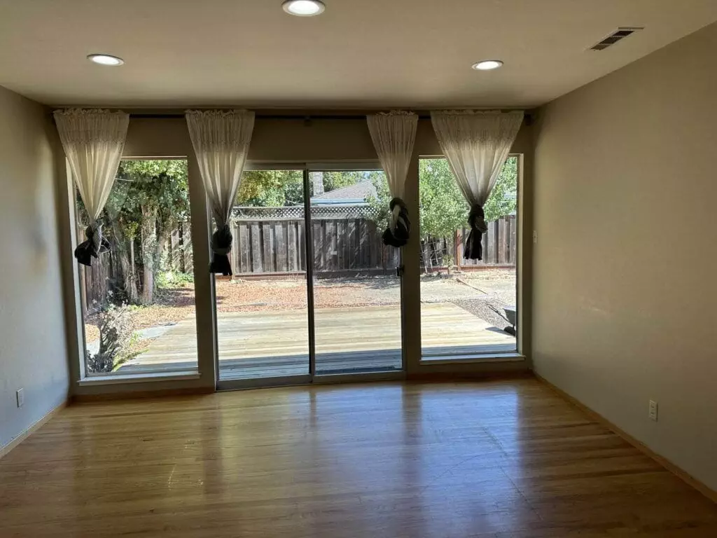 An empty room with wooden flooring and sliding glass doors leading to a wooden deck and backyard. White curtains are tied back, and there are recessed lights on the ceiling.