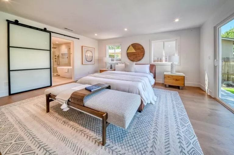 Contemporary bedroom with a large bed, wooden nightstands, a cushioned bench, a sliding door to an ensuite bathroom, a rug, and two large round wall mirrors. Ample natural light from windows.