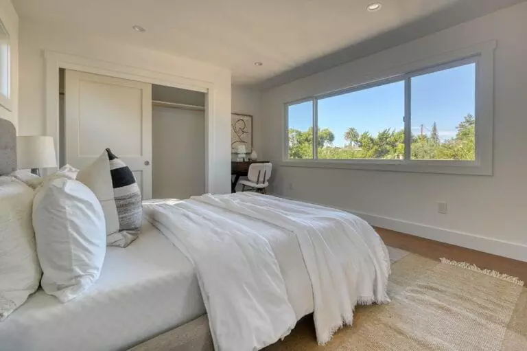 A bright bedroom with a neatly made bed, white bedding, cushions, an open closet with white sliding doors, and a large window overlooking greenery.