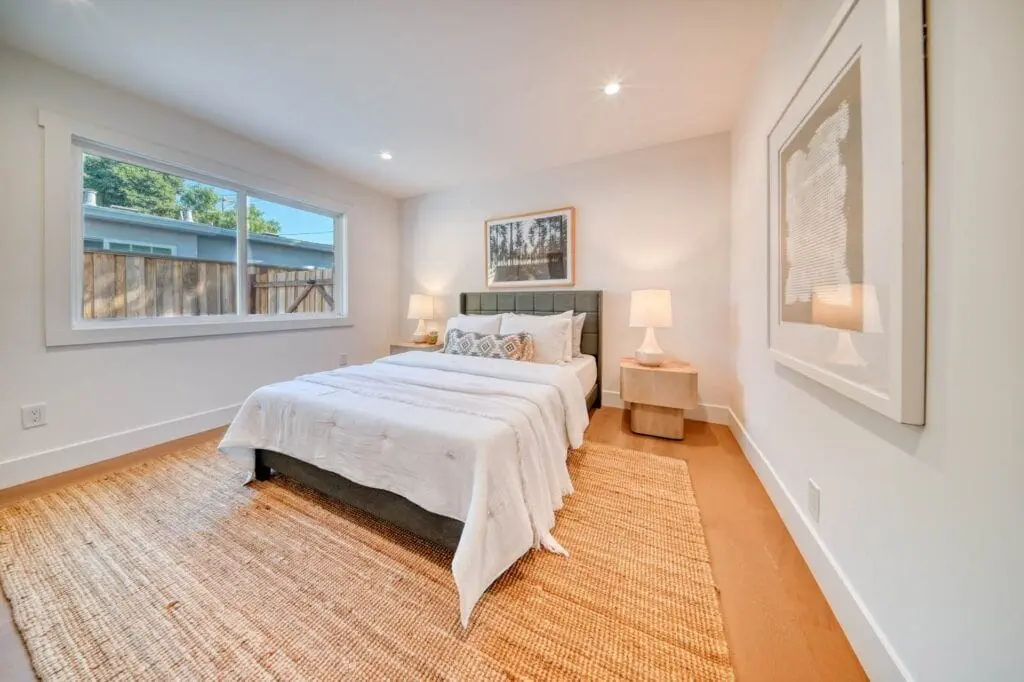 A minimalist bedroom with a double bed, white linens, two bedside lamps on wooden tables, a large window, and framed wall art. The room has wooden flooring and a woven rug.