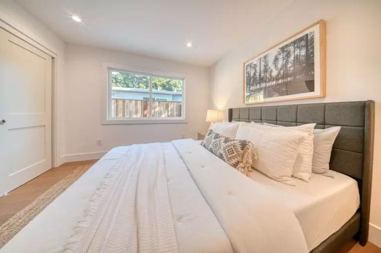 A modern bedroom with a large bed, white bedding, a padded headboard, a bedside lamp, and a large window. A landscape artwork hangs above the bed.