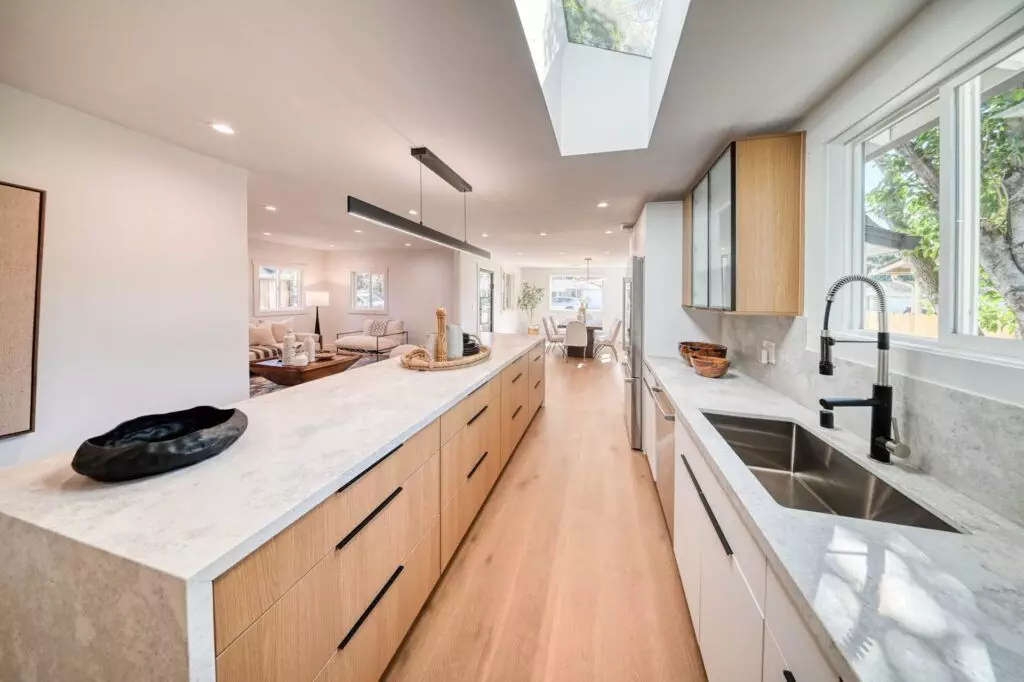 Modern kitchen with a long white marble countertop, wooden cabinets, stainless steel sink, and overhead skylight. An open living and dining area is visible in the background.