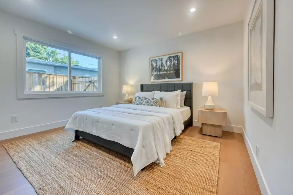 A modern bedroom features a neatly made bed with white linens, two bedside tables with lamps, a large window, a woven rug, and framed artwork on the walls.