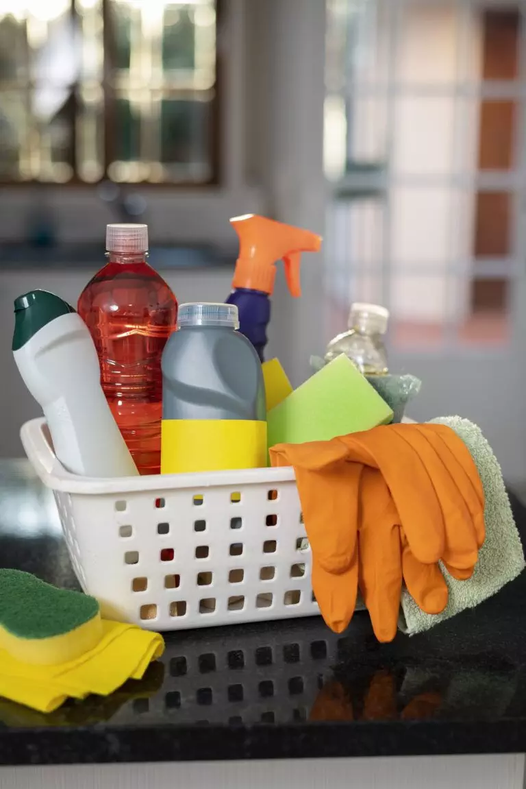 A white basket containing various cleaning supplies, including spray bottles, sponges, rubber gloves, and cleaning cloths, sits on a kitchen counter in a Santa Clara home.