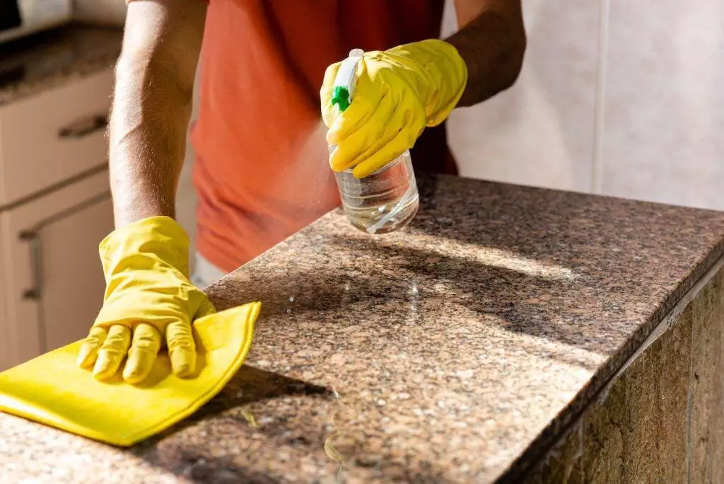 In a recent ApartmentGuide article, a person wearing yellow gloves meticulously cleans a granite countertop using a spray bottle and cloth.